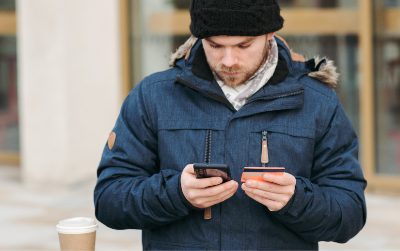 Man in winter coat and hat using a smartphone and holding a credit card, potentially shopping for trending Amazon products.